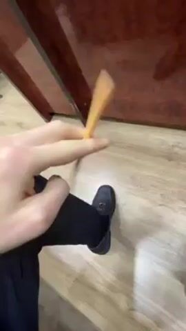A person wearing navy blue pants and dark blue shoes sits indoors on a light wood floor next to a dark brown table with a shiny surface. The person holds a yellow pencil in their right hand and twists it back and forth.