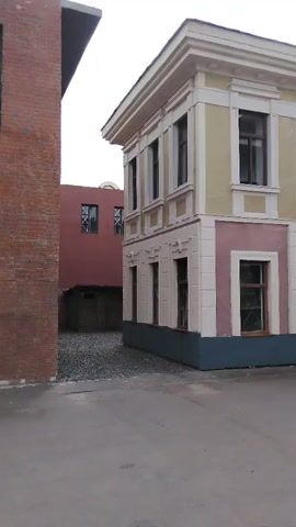 A handheld camera pans slowly to the right, showing the exterior of two buildings. The building in the foreground is a two-story structure with a pale yellow upper floor and a pinkish-brown lower floor. It has white trim and dark-framed windows. The building on the left is a red brick structure, and partially obscures a smaller, dark red building behind it. This building has two dark-framed windows. The ground in the foreground is asphalt, and the ground between the buildings is covered in dark gravel. The sky is overcast.