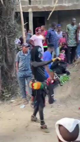 In an outdoor setting, a video captures a community gathering with several individuals dressed in various traditional costumes. The costumes range from vibrant red and yellow outfits with intricate headwear to black and white ensembles reminiscent of pop culture figures. They are surrounded by a crowd of people, some casually dressed, observing the costumed individuals. The environment appears to be a village or rural area with sandy ground and scattered vegetation, including palm trees. Motorcycles are parked in the background, suggesting a mix of traditional and modern elements in the setting. The overall atmosphere seems celebratory, potentially indicating a local festival or cultural event.