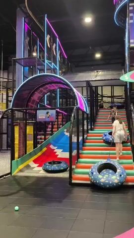 The video pans across a vibrant indoor playground filled with children and adults. A girl in a white top and grey shorts ascends a colorful staircase, holding an inner tube. In the foreground, two girls sit on a pink bench, one in a white top and grey pants, the other in a black t-shirt and shorts. Behind them, a ball pit is visible with several children playing. Further back, a large colorful circular structure with an orange and blue design dominates the space, with a yellow pole structure to its right. Neon lights and playful signage, including the word "HELLO," add to the energetic atmosphere of the playground. The overall environment is that of a modern, brightly lit entertainment facility.