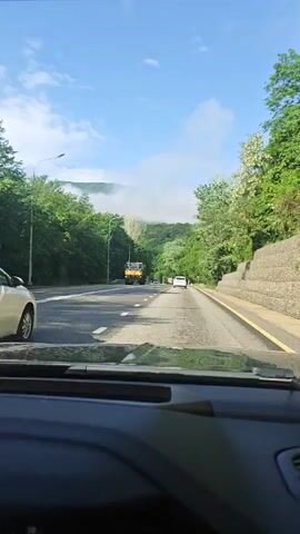 A camera, possibly mounted in a vehicle, records a drive on a road with trees on both sides. A light blue sky with sparse clouds is visible. There are other vehicles on the road, including a white car in front of the camera's vehicle. A hill shrouded in fog or low clouds is ahead. The road has white lane markers.