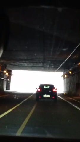 The video begins inside a vehicle driving through a tunnel. The tunnel is dark with bright light visible at the end. A black SUV is in front of the vehicle. As the vehicle exits the tunnel, the view opens up to a mountainous landscape. The road is a highway with multiple lanes. The same blue SUV is still in front. The weather appears to be overcast and rainy, with water droplets visible on the windshield. The mountains are steep and rocky with patches of vegetation. Road signs are visible along the highway, indicating directions and potential hazards. The vehicle continues on the highway, following the blue SUV.
