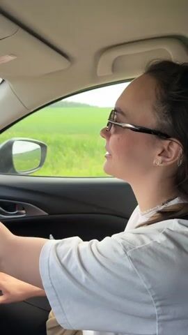 The video captures a woman driving a car during the daytime, likely in a rural area like Lukintsy, Russia.

**Subjects:** The primary subject is a woman, presumably the driver, seen from the perspective of a passenger. She has dark hair, wears black-framed glasses, and a white short-sleeved t-shirt. She has light freckles on her nose and cheeks and a serene expression, looking straight ahead. Her right hand is visible on the black steering wheel, adorned with a silver-toned rectangular wristwatch with a metal band.

**Scene and Setting:** The interior of the car is black. The external view through the windows and windshield shows vast, vibrant green fields under a bright, possibly overcast sky, suggesting open countryside. The road is visible at the bottom of the windshield in one shot.

**Activity or Event:** The woman is driving the car, navigating what appears to be a rural road. The overall activity is a casual drive.

**Notable Details:**
*   A red, oval-shaped air freshener with white text (partially obscured, possibly "AROMATIKA") hangs from the rearview mirror.
*   Several sticky notes in yellow, pink, and green are affixed to the dashboard and possibly the lower part of the windshield.
*   A small electronic screen on the dashboard displays "Bluetooth Ольга" (Bluetooth Olga) and "Между строчек" (Between the lines), indicating a connected device or current audio playback, and also serving as a location cue suggesting a Russian-speaking environment.
*   A dashcam or similar device is mounted at the top center of the windshield.
*   The time of day is bright daylight, and the weather appears clear or lightly overcast.