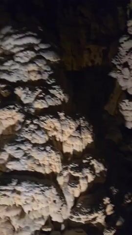 The video shows an interior view of a cave with stalactites and stalagmites. The camera pans slowly, illuminating the rock formations with artificial light. The cave walls and ceiling are visible, as are numerous speleothems of varying sizes and shapes. The lighting casts shadows, creating a dramatic effect. The environment is dark and cavernous, with the formations appearing to be made of stone. There are no people or explicit objects other than the cave's natural formations. The video appears to be a guided tour or exploration of the cave.