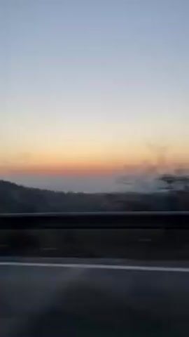 The video shows a sunset view from a moving vehicle, likely on a highway. The sky is transitioning from a light blue to orange and pink hues near the horizon. A silhouetted landscape with trees and distant mountains is visible beneath the colorful sky. A metal guardrail is visible in the foreground, with the road surface below. The camera appears to be inside a car, capturing the scenery through the window.