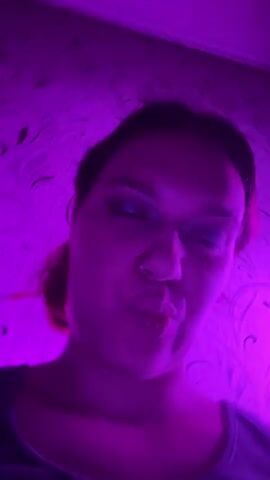 A person's face is illuminated by a purple light source that creates a halo effect around their head. The person is looking at the camera and their tongue is sticking out. The background is a textured wall with a swirling pattern. A circular light fixture, likely a ring light, is visible in the lower right corner of the frame. The lighting is dramatic and gives the scene a vibrant, almost otherworldly feel. The video is very short, lasting only a few seconds.