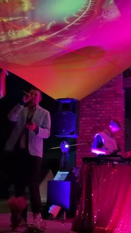 The video captures a live performance at night, possibly outdoors or in a venue with an open ceiling covered by a draped fabric illuminated with colorful projections of yellow, orange, and green. The stage is set with musical instruments and lighting. A man in a light-colored jacket and dark pants stands center-left, holding a microphone and singing. To his right, a person in a vest is seated behind a keyboard, playing the instrument. The keyboard is covered with a sequined red fabric. Further to the right, a woman in a vibrantly patterned outfit sings into a microphone and plays an acoustic guitar. Next to her, a man with a bald head and wearing a blue jacket plays an acoustic guitar. He appears to be singing as well. In the background, a brick wall is visible, and an upper level with railings and people looking down can be seen. The overall lighting is dominated by pink and purple hues, with blue lights also present. The environment suggests a festive or artistic gathering.