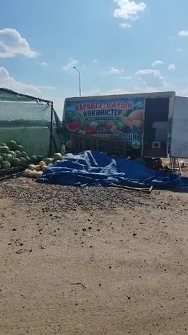 The video shows an outdoor market stall selling fruits and vegetables under a bright blue sky with scattered clouds. In the foreground, there is a dusty, gravelly ground. To the left, a large quantity of watermelons is visible, some stacked under a green mesh canopy. In front of the watermelons, a pile of lighter-colored melons, possibly cantaloupes or honeydew, is arranged. A set of scales is also present near the watermelons. Dominating the center and right of the frame is a white truck or trailer with an open side, serving as a display area. A large blue tarp is spread out in front of the truck, partially covering some produce. A sign with colorful images of fruits and vegetables and text in a foreign language is attached to the truck. Various fruits and vegetables are visible on display, including watermelons, pumpkins, and possibly other gourds. A small table and stool are located to the right of the truck. The scene appears to be a roadside stand, likely in a rural or semi-rural area. The lighting suggests it is daytime, with the sun casting shadows.