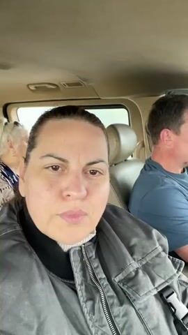 Inside a tan-colored vehicle, a woman in a gray jacket takes a selfie video. The driver, a man in a blue shirt, glances out the window. The woman looks directly at the camera, and the man rubs his face. Greenery is visible through the window. The interior of the vehicle is clean and well-maintained.