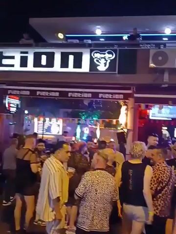 The video shows a crowded street at night, lined with bars and restaurants. People are walking, standing, drinking, and talking. The signs for the bars "Fiction" and "Mardi Gras" are visible. The lighting is bright and festive. The atmosphere appears lively and social.