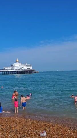 The video shows a wide shot of a beach on a sunny day. The water is a clear blue and there are several people in it, some swimming and some wading near the shore. In the background, a white pier with a dome-shaped structure on it extends out into the sea. The beach itself is made of pebbles.