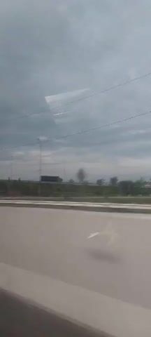 The video shows a view from inside a vehicle looking out at a highway scene. The perspective is from a side window, with reflections visible on the glass. The time stamp indicates the video starts at 00:00 and ends at 00:05.

In the background, a cloudy sky with some wispy clouds is visible. Below the sky, there are power lines and streetlights. Further down, a road can be seen with a dark-colored truck driving on it. There are trees and vegetation along the side of the road.

As the video progresses, the vehicle appears to be passing under an overpass or a bridge structure. The underside of the overpass is visible, with its concrete supports. The landscape continues to be a mix of trees and open areas. The foreground consists of the interior of the vehicle, with the window and reflections creating a sense of motion. There are no people visible in the video. The overall environment is that of a typical highway setting, possibly in a suburban or rural area.