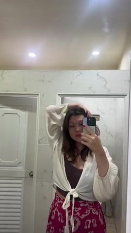 In a bathroom, a young woman stands in front of a mirror, holding up her smartphone to take a selfie. She has brown hair, a pearl necklace, a brown top, a white button-down shirt tied at the waist, and a red floral-patterned skirt. She throws her hair, smiles, and sticks her tongue out at the camera, before making an exaggerated gasp. The bathroom has white marble-patterned walls, two doors, and two ceiling lights.