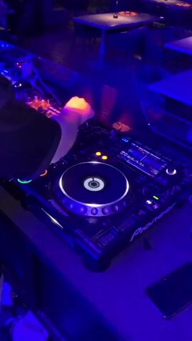 A close-up, high-angle shot shows a person's hands operating a DJ setup in a dimly lit establishment. The DJ equipment, primarily a Pioneer CDJ-2000 nexus, is the central focus. The DJ's right hand is seen adjusting knobs on the mixer, while their left hand hovers over the jog wheel of the CDJ. The equipment features illuminated buttons and displays, indicative of its active use. The background reveals a blurred view of tables and chairs, suggesting a lounge or club environment, with ambient blue and purple lighting casting a glow. A smartphone lies on the surface in the foreground.