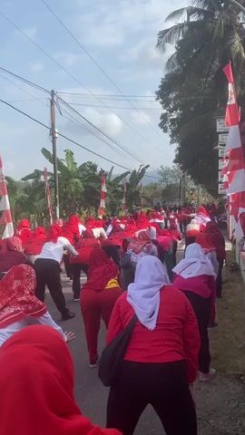 A group of people, predominantly women wearing red clothing and headscarves, are performing stretches outdoors on a paved road. They are arranged in rows, facing away from the camera and bending over. The scene is decorated with numerous red and white flags, likely national flags, spaced along the roadside. Banana trees and other foliage are visible in the background, suggesting a rural or semi-rural setting. The sky is bright, indicating daytime. The camera is positioned behind the group, looking down the road at them. The audio contains music.