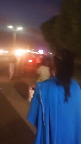 At night, a person wearing a blue garment films the scene in front of them. There are people walking on the street and sidewalk in the distance. Headlights from cars illuminate the area. A street light shines brightly. A tree is visible on the right. Someone is heard speaking in French. The scene is blurry and the camera appears to be in motion.