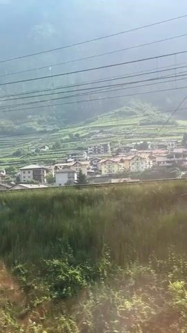 The video captures a train's view of a village nestled in a valley, with terraced vineyards covering the hillsides leading up to a misty mountain range. Power lines and poles are visible in the foreground and mid-ground, suggesting the train is on an electrified route. The sun appears to be shining, creating a bright sky with scattered clouds. The architecture of the village consists of multi-story buildings with tiled roofs, suggesting a European setting. The camera is moving, indicating the video was filmed from a moving vehicle, likely the train.