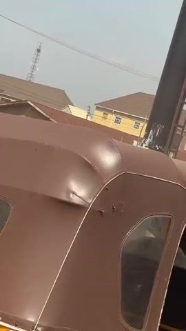 The video captures a street scene in Katambe, Nigeria, during the daytime under a bright, clear sky.

Initially, several yellow Keke Napeps (auto-rickshaws) are visible, parked or driving on an unpaved, reddish-brown dirt road. Buildings with varying roof colors and a distant communication tower stand in the background.

The camera then shifts to a large, green and red truck moving past. The truck's cargo area is covered with a green tarp, secured with ropes. A person's bare legs are briefly visible on top of the tarp, indicating someone is riding on the truck's roof or cargo. As the camera pans up, a tall, black pole with a ladder attached is seen, with part of the truck and the person on top still in view. The video concludes with a shot of the bright sky, featuring power lines and a billboard structure.