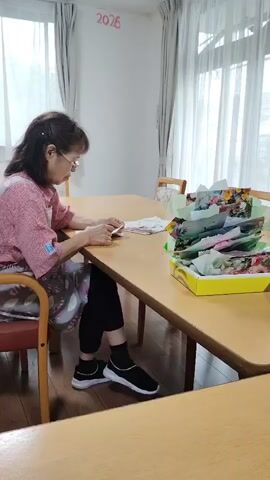 A static shot shows two elderly women sitting at a wooden table in a room with large windows and curtains. The woman on the left, wearing glasses and a pink apron over a patterned shirt, is facing the camera and looking down at something in her hands. She has dark hair and is wearing black pants and sneakers. A box filled with colorful paper and decorative items sits on the table between the two women. The woman on the right, with gray hair, is facing away from the camera, looking down at her hands and working on something on the table. She is wearing a dark brown shirt. There are drawings or decorations on the wall behind the woman on the right. The number "2026" is written in red on the wall above and to the left of the windows. The lighting suggests an indoor setting, possibly a common room in a care facility or community center.