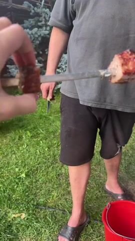 The video begins with a close-up of a hand holding a skewer with a piece of meat on it. The skewer is being held in front of a person wearing a gray t-shirt and black shorts. The person is standing in a grassy area with a red bucket at their feet. The skewer appears to be metal. The hand holding the skewer is about to remove a piece of meat from it. The camera then pans to the right, showing a blurred background of trees and a building. The video ends with a shot of a table with plates, glasses, and a faucet. The table appears to be outdoors.