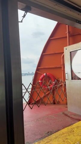 The video captures an outdoor view from what appears to be the deck of a ferry. The foreground shows a red deck with yellow markings along the edge. A collapsible metal gate, expanded to form a barrier, is positioned in the middle ground. Behind the gate, an orange wall of the ferry rises diagonally, and a red life preserver with white stripes is attached to it. To the right, a white door with a small circular window is visible. Beyond the ferry's structure, a body of water stretches out, with a distant cityscape visible on the horizon under an overcast sky. The overall impression is one of being on a moving vessel, with the vastness of the water and the city in the distance.