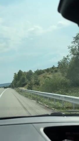 The video is a first-person perspective from inside a vehicle, looking out the windshield. The vehicle is traveling on a paved road with a white line marking the lane. To the left of the road, there is a grassy shoulder and a dense line of green trees and bushes. To the right of the road, there is a hill covered in grass and scattered trees, with a metal guardrail separating the road from the embankment. The sky is cloudy and blue. The video captures the motion of the vehicle as it travels along the road, with the scenery passing by.