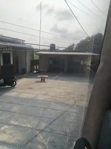 The video shows a view from inside a vehicle, looking out through a window. The exterior scene is somewhat blurry and the lighting suggests it is daytime. In the foreground, the side of a vehicle seat is visible. The exterior shows a paved area with square tiles, a covered structure with a corrugated roof, and some trees and vegetation. There is a water tank on top of the covered structure. Further back, there are more trees and what appears to be a dirt road. In the last few seconds, a string of colorful flags is visible. There are no people clearly visible in the footage. The overall impression is of a rural or semi-rural environment.