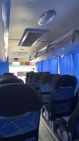 This media file depicts the interior of a bus with rows of empty seats. The seats are upholstered in dark fabric with a blue and yellow patterned fabric on the lower portion. Blue curtains line the windows on the right side of the bus. The ceiling is white and features overhead lighting and an air conditioning unit. A digital display showing "00:00" is visible at the front of the bus, suggesting it might be a clock or timer. Signs with red and yellow elements are also visible at the front, though the text is not legible. The overall scene suggests the bus is either at the beginning of a journey or has just completed one, as the seats are unoccupied. The lighting indicates it is daytime, and the interior of the bus appears clean and well-maintained. There are no people visible in the frame, reinforcing the idea of an empty bus. The visual style of the video is a first-person perspective, likely from someone walking down the aisle of the bus.