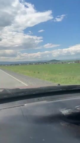 This video captures a view from inside a vehicle, looking forward through the windshield. The vehicle appears to be moving along a paved road, bordered by a gravel shoulder on the left. To the right of the road is a vast expanse of green farmland, with a line of houses visible in the distance, forming a village or town. The sky above is a vibrant blue, dotted with fluffy white clouds. The foreground is dominated by the dark interior of the car's dashboard. The camera appears to be held steady, suggesting it might be mounted or the driver is being careful. The overall impression is one of a scenic drive through a rural landscape on a bright, partly cloudy day.