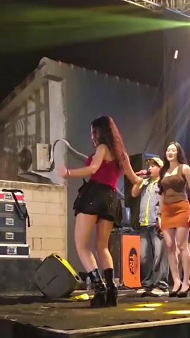 A young woman with long dark hair, wearing a red strapless top and a black sequined skirt, dances energetically on a stage. She is wearing tall black platform boots. To her right, another young woman in a brown top and orange skirt stands observing. Behind them, there are speakers, equipment cases, and stage lighting that casts colorful hues. The background appears to be a makeshift set resembling a building facade. The scene captures a live performance with a focus on the central dancer's dynamic movements.