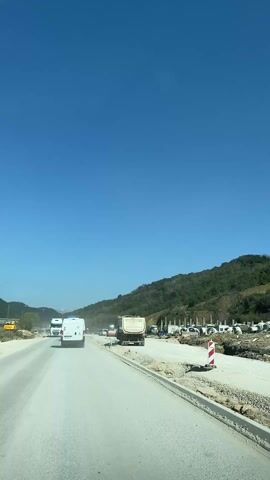 The camera is mounted inside a vehicle moving forward on a road under a clear blue sky. Several other vehicles, including a white van and a large truck, are also driving in the same direction. To the right of the road, there is a construction area. Large cylindrical concrete pieces are stacked in the area. A hill covered in green vegetation rises in the background. The right side of the road is under construction, with a partially paved section and exposed gravel. A red and white striped traffic marker stands near the construction. The scene appears to be a highway undergoing roadwork on a sunny day.