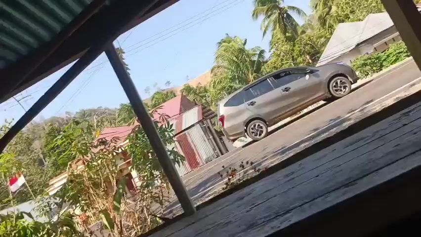 The video shows a residential street with a gray car driving away from the camera. The street is lined with palm trees and houses. The video appears to be filmed from the porch of a building. The building has a corrugated metal roof and wooden supports. The porch also has wooden floorboards. In the background, there are hills covered with trees and vegetation. The sky is blue and clear, indicating a sunny day.