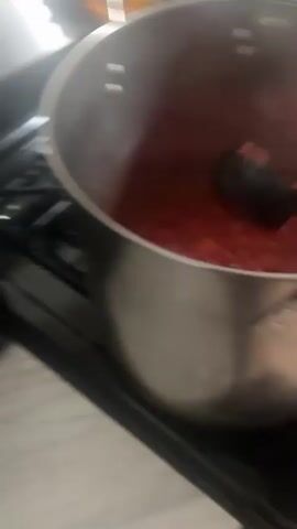 A red, lumpy sauce is cooking in a large, silver pot on a stovetop. A black spoon or spatula is visible, occasionally stirring the sauce. The camera angle suggests the person holding it is likely cooking. Other kitchen items can be seen blurred in the background.