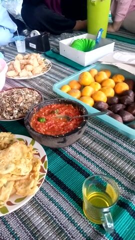 The image depicts an overhead view of a picnic or meal laid out on a woven mat. The scene features a variety of Indonesian dishes. In the center, a mortar and pestle filled with a vibrant red sambal, likely a chili paste, is prominent. To its left, a bowl contains what appears to be fried fish or crackers, and a pink bowl holds white rice. Next to the sambal is a shallow dish filled with dried anchovies or small dried fish. To the right, a plate overflows with golden-brown fried tempeh.

Further back, a light blue plastic container holds a bounty of ripe oranges and dark, scaly salak (snake fruit). A glass of yellow liquid, possibly tea or juice, sits to the right of the food spread. In the upper left, a bright green, circular object, possibly a decorative item or part of a food presentation, is visible next to a white box. The lower left corner shows a person's legs and arm, indicating their presence at the meal. The overall setting suggests an informal outdoor gathering. The lighting is bright, hinting at daytime, and the food itself suggests a meal being shared. The presence of fresh fruit and traditional Indonesian dishes points to a meal enjoyed in Indonesia, possibly in a setting like Sukaraja. There is no visible text in the image.