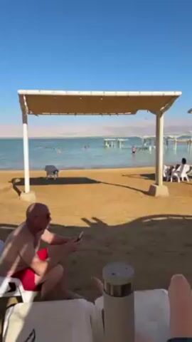 The video shows a view of the Dead Sea on a sunny day. The camera pans from right to left across a sandy beach with several people relaxing under white-roofed sunshades. In the water, several people are floating. The water is a calm, turquoise color, and there are mountains visible in the distance across the sea. A man in red swim trunks is seen looking at his phone. A woman and a young boy walk along the beach.