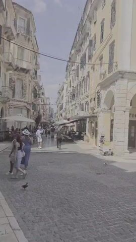 The video pans across a cobblestone street in a European city, featuring old buildings with balconies and shutters. Pedestrians walk along the street, and some shops appear to be open. A few pigeons are visible on the ground. The scene transitions to a wider view showing a plaza area with trees and more buildings. Several vehicles, including cars and a van, are parked or driving in the area. The sky is partly cloudy.