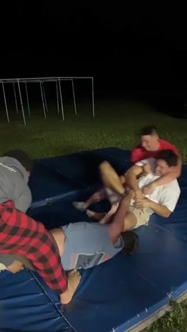 The video depicts four young men engaged in a playful wrestling match on a blue mat outdoors at night. The scene is illuminated by artificial light, casting shadows and highlighting the subjects. One person, wearing a grey hooded sweatshirt and red and black plaid pajama bottoms, is on the left, appearing to initiate a move. Another individual in a white t-shirt and khaki shorts is on his back, laughing heartily. A third person in a red t-shirt is lying on top of the laughing individual, also seemingly participating in the fun. The fourth person, wearing a blue t-shirt, is on the left, facing away from the camera and appears to be on the ground.

The setting is a grassy area, with what looks like a metal frame structure in the background and a dark car parked behind it. The overall atmosphere is one of lighthearted camaraderie and physical exertion. The individuals' expressions suggest amusement and enjoyment of the activity. The time of day is clearly night, given the dark background. There are no discernible weather conditions, but the grass is green, indicating it's likely not winter. There is a license plate on the car in the background, but the text is not legible.
