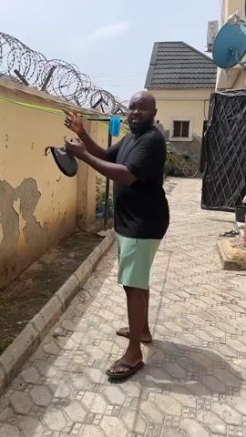 The video shows a man standing in a courtyard and gesturing to the camera. The man has a beard and is wearing a black shirt, green shorts, and sandals. He is standing on a patterned tile surface.  Behind him is a wall topped with barbed wire. There is a building in the background with satellite dishes on the roof. The man walks toward the camera. The camera then shows a close-up of a patched area on the wall near a window.  A barrel sits below the wall.