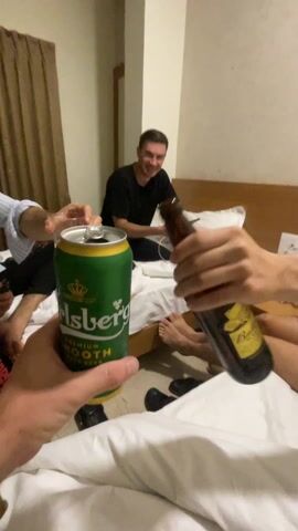 Three men are in a hotel room. Two are sitting in the bed, and one is at the end of the bed. They are toasting drinks, a glass of amber liquid, a can of Carlsberg beer, and a bottle of beer. There is a wooden bedside table with a phone and some remotes. The room has a white wall and a tan curtain.