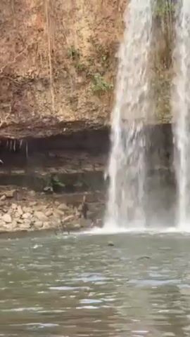 A full shot depicts a waterfall cascading into a body of water. The cliff face beside the waterfall is layered with light brown rock, with some greenery sprouting in the crevices. A thin figure is visible in dark clothing beside the waterfall, appearing to move or adjust their position on the rocks near the water. The water below the falls appears dark and turbulent, reflecting the surroundings. The light is bright, illuminating the water and the waterfall, which creates a diffuse, white spray.