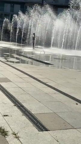 The video shows a person standing in the middle of a dry fountain, with water jets spraying up around them. The fountain is made of light-colored paving stones with metal grates for drainage. The person is wearing dark clothing. In the background, there is a building with multiple stories and windows, and some greenery. The sun is shining, casting shadows on the ground.