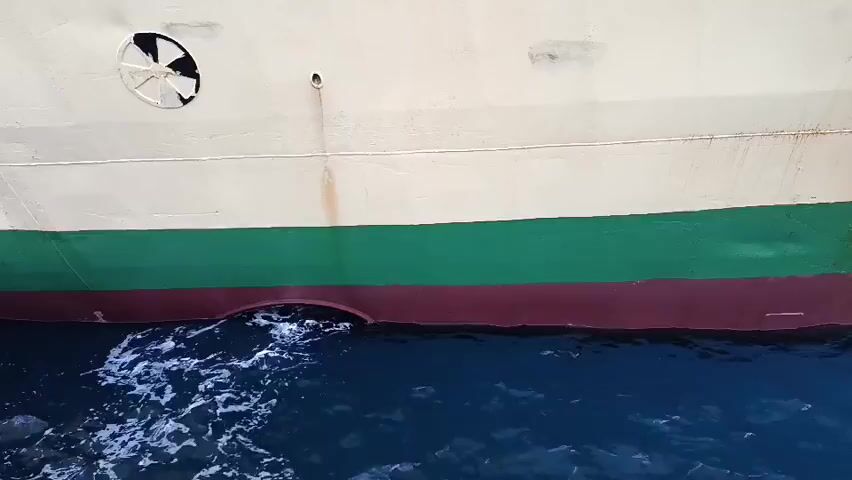 A video clip shows the side of a ship in dark blue water. The ship's hull is painted white, with a green band and a maroon band visible. There is a circular, fan-like object with broken blades on the white part of the hull. Below the maroon band, water is churning and splashing, indicating the ship is in motion or a propeller is active. A thin stream of water is seen spraying from a small opening on the side of the hull, just above the churning water. The water is choppy and creates a foamy wake as it interacts with the hull. The overall scene is one of a vessel moving through the sea.
