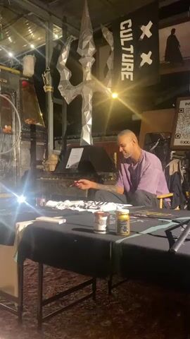 A middle-aged man with a goatee and a purple T-shirt sits at a black table indoors, working with a paintbrush on a white cloth with dark designs on it. Two jars of paint are in front of him on the table. Above him is a silver, geometric trident-shaped sculpture and a flag that says "CULTURE." A bright light is shining over the table. There is a sign visible behind the man that says "STELLA."