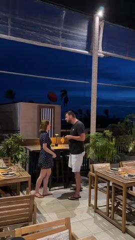 A man and a woman stand facing each other at a table on an outdoor patio restaurant at dusk. They both hold drinks. The patio is filled with tables and chairs, some occupied by other diners. The sky is dark blue with some clouds. There are plants and lighting fixtures on the patio.