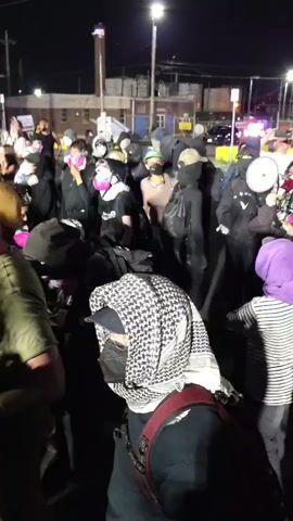 A large group of people is gathered outdoors at night in an urban environment in Newark, USA. The scene is illuminated by streetlights, with various buildings visible in the background, suggesting an industrial or commercial area. Distant flashing lights, possibly from emergency vehicles, can be seen further down the street.

Many individuals in the crowd are wearing face coverings, including balaclavas, medical masks, and keffiyehs, with several also sporting respirators or gas masks. People are dressed in casual attire, such as jackets, hoodies, and t-shirts, and some are carrying backpacks.

The group appears to be engaged in a protest or demonstration. One person is speaking into a megaphone, leading chants. The most clearly audible chant is "Lady Hall must fall." Participants are actively responding to the speaker. A person prominently featured in the foreground, seen from behind, is wearing a black and white keffiyeh. Partial text, "BLACK," is visible on the shirt of one participant. The atmosphere conveys a sense of collective action and determination.