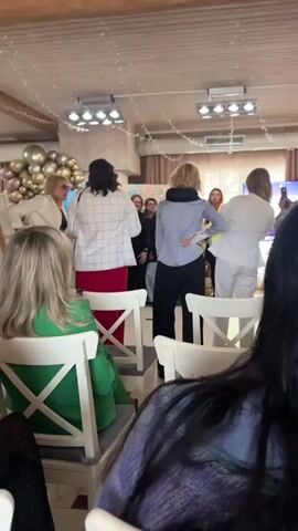 The video shows a gathering of people in a brightly lit, festive indoor setting. Most of them are standing and seem to be posing for a photo or watching some kind of performance or event. There is a balloon arrangement in the background. The room has a wooden ceiling decorated with string lights. Several people are seated in chairs in the foreground, watching the scene. Some individuals are dressed in white, while others wear various colors. A man in a suit is taking a photo with his phone. The overall atmosphere appears cheerful and celebratory.