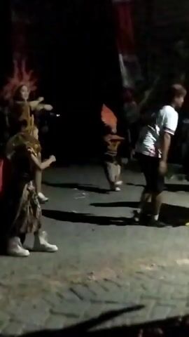 The video shows an outdoor nighttime event. In the foreground, a child wearing an elaborate costume with a gold headdress and a patterned skirt stands on a paved surface. The child is holding a white placard with the number "08" written in black. To the right of the child, a person in a white t-shirt and dark shorts is visible. In the background, other people are present, some wearing costumes, and there appears to be some lighting and decorations, possibly flags, further in the background. The scene suggests a parade, festival, or competition.