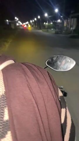 The video shows a first-person perspective of someone riding a motorcycle at night. The rider is wearing a brown sweater with a patterned design. The road is lit by streetlights and the headlights of oncoming vehicles. The surroundings are blurred due to the motion of the motorcycle. The camera is focused on the rider's shoulder and the road ahead.