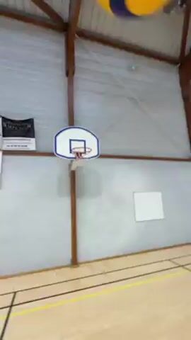 A video shows someone bouncing a yellow volleyball from a basketball hoop. The camera is held from the first person point of view, showing only the arm of the person bouncing the ball. The volleyball bounces once off the rim of the hoop and then hits the floor, rolling out of the frame. The video is shot in an indoor gymnasium with a basketball hoop attached to a wall. The wall is gray and white and has a white square whiteboard. Yellow lines mark the floor.