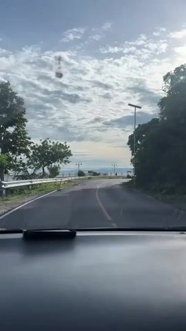 The video shows a view from inside a car driving on a winding road. The road is paved with asphalt and has a yellow line down the center. On the left side of the road, there is a white railing and trees. On the right side of the road, there are trees and a solar-powered lamp post. In the distance, there is a body of water and a cloudy sky with the sun shining through. The car appears to be moving slowly.